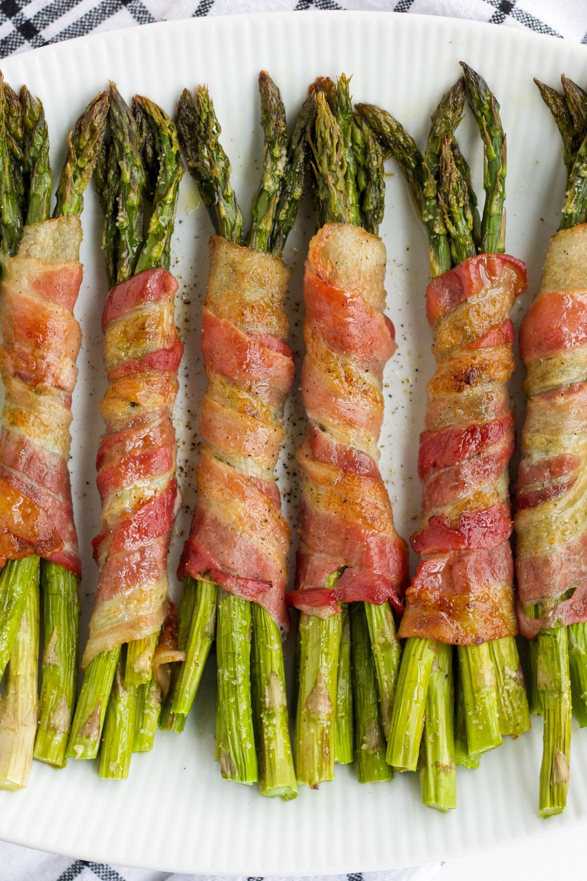 Six portions of asparagus wrapped in bacon on a white plate.
