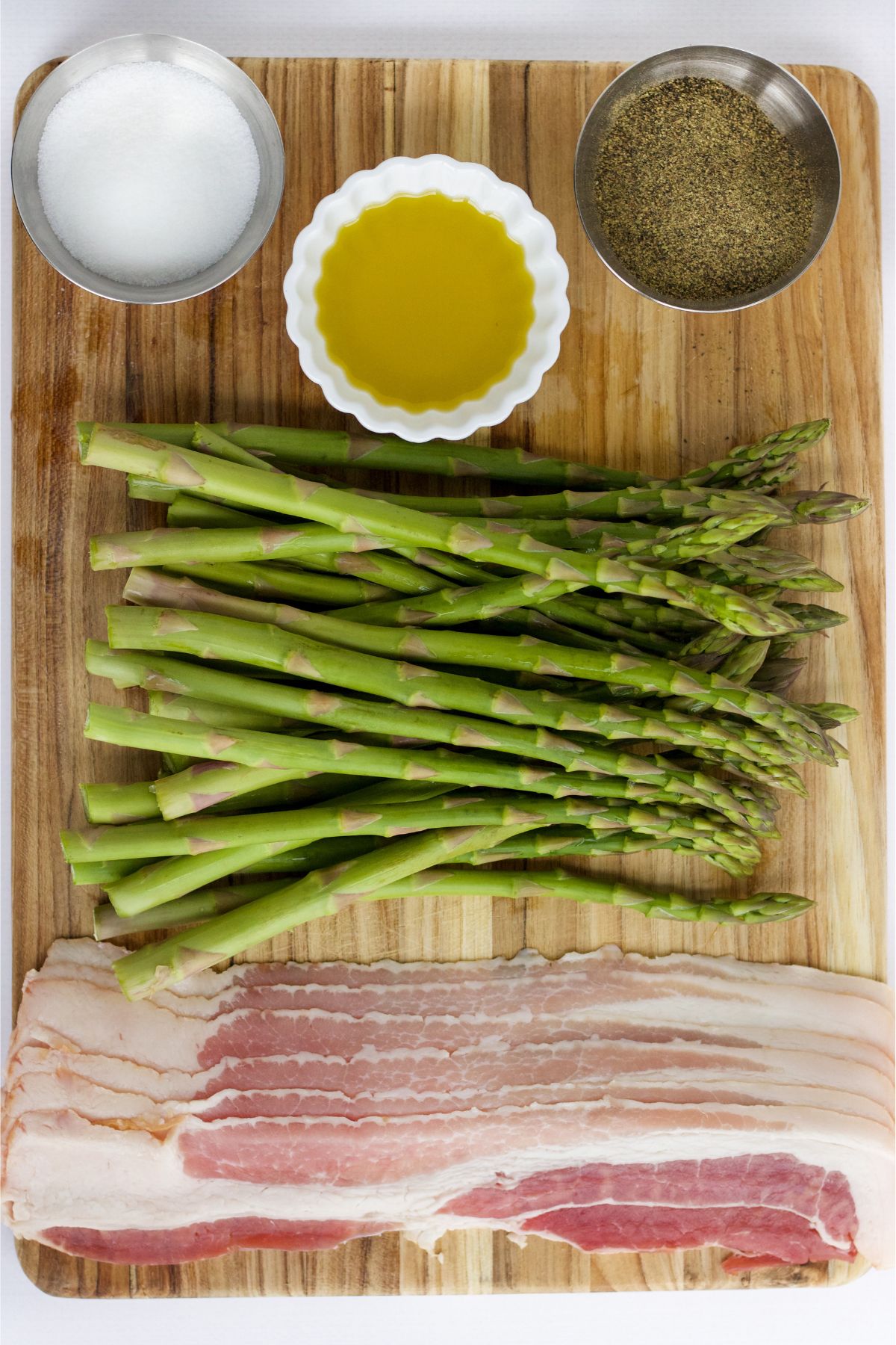 The ingredients for asparagus wrapped in bacon on a cutting board.