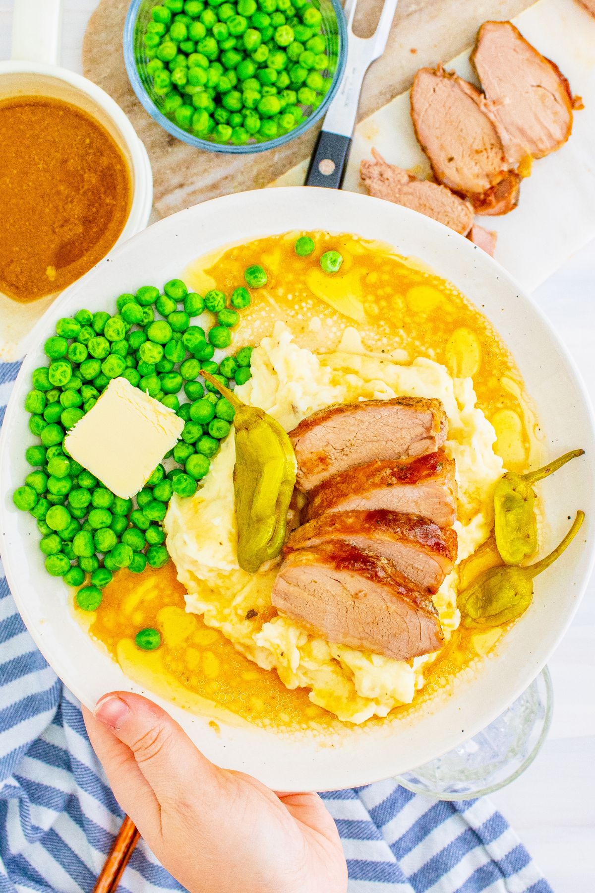 A white plate with pork tenderloin over top of mashed potatoes and a side of peas.