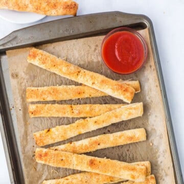 Pizza dough breadsticks on a sheet pan with a side of marinara sauce.
