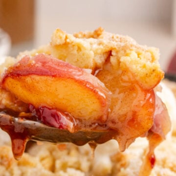 A fork holding a bite of Grandma's Peach Cobbler with a crispy golden crust, dripping with peach filling, and a scoop of vanilla ice cream in the background.