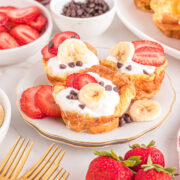 A plate with three German pancake muffins topped with yogurt, sliced strawberries, bananas, and chocolate chips.
