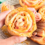 A close-up of a hand holding a ham and cheese pinwheel with other pinwheels visible on a plate in the background.