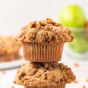Two apple crisp muffins stacked on top of each other with green apples in the background.