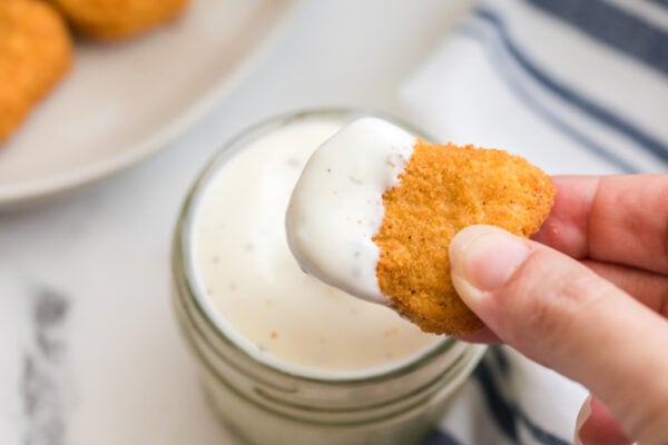 A hand dips a breaded Air Fryer Chicken Nugget into a jar of white dipping sauce, with a striped cloth in the background.
