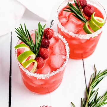 Two glasses of cranberry margarita garnished with rosemary, cranberries, and lime.