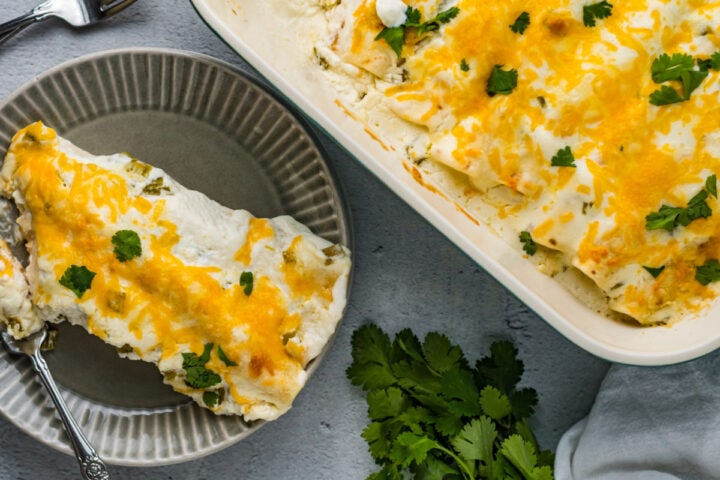 Overhead photo of Sour Cream Chicken Enchilada served in a small gray-colored plate with a fork. More Enchilada in a large-rectangular-white-colored dish. The enchiladas are garnished with chopped cilantro.