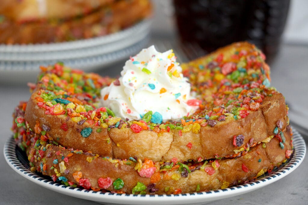 plate of two slices of air fryer fruity pebble french toast topped with whipped cream. More Fruity Pebble French Toast and a glass container full of syrup in the background.