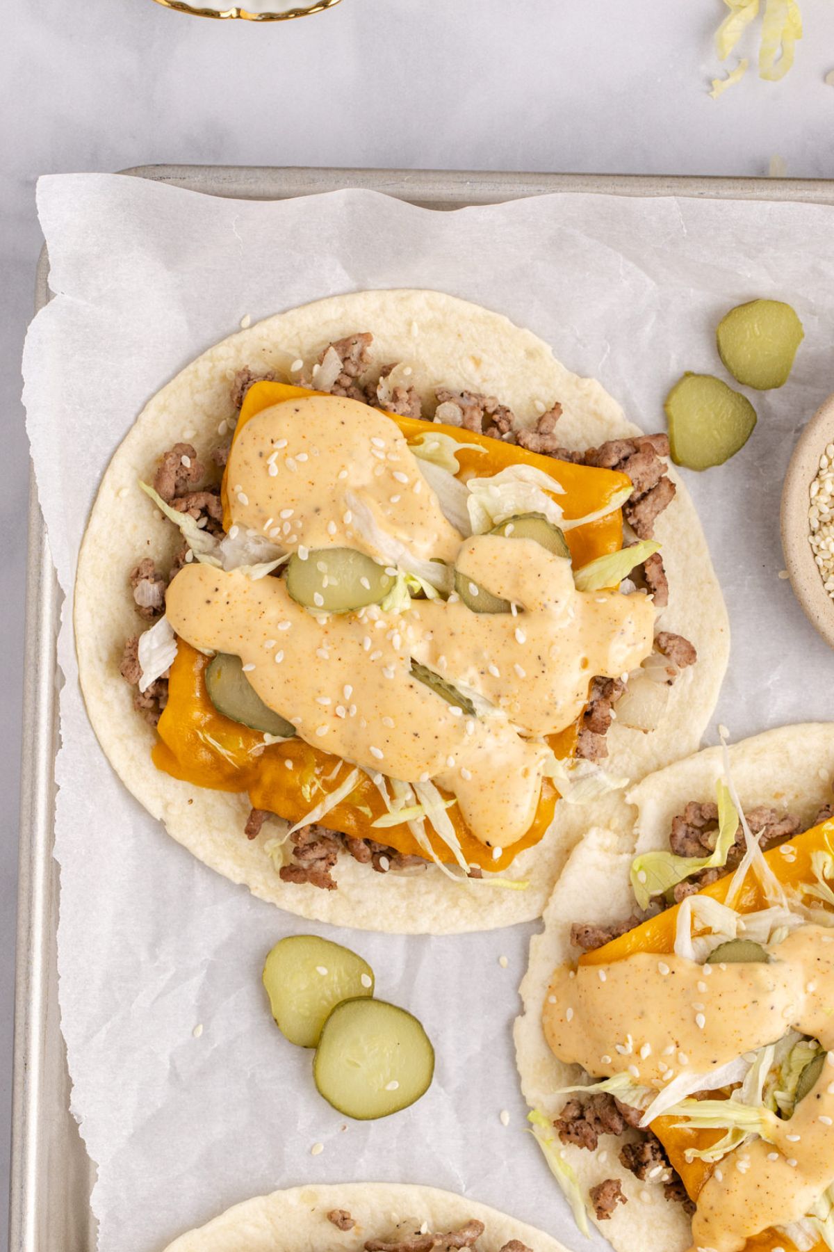 A sheet pan of burger tacos with cheese, pickle slices, lettuce, and sauce over beef.