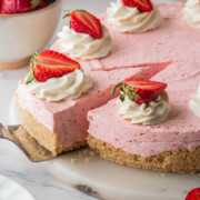 A spatula lifting a slice of no-bake strawberry cheesecake with whipped cream and strawberries on top.