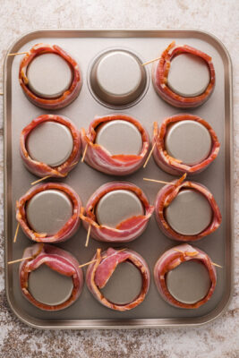 Bacon wrapped around the bottom of a muffin tin.The bacon is secured using toothpicks.