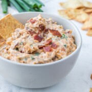 A bowl of creamy dip garnished with bacon bits, accompanied by wheat crackers.