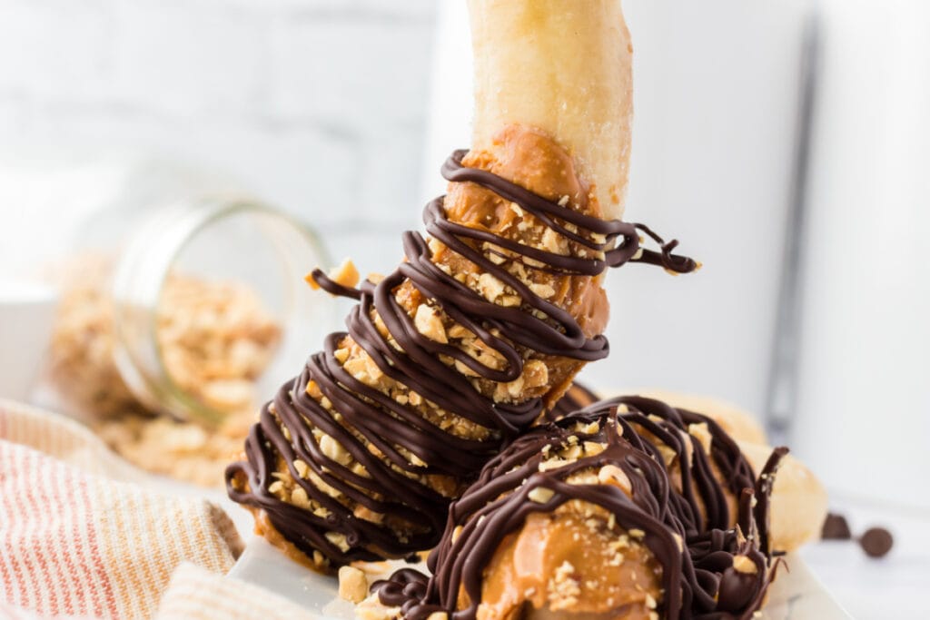 Two peanut butter chocolate bananas on towel. The bananas are coated in peanut butter and crushed peanuts. Chocolate is spread over the banana like a string giving the illusion it is binding the peanut butter and banana together. One Peanut Butter Chocolate Banana is standing upright and leaning against another which is lying down.