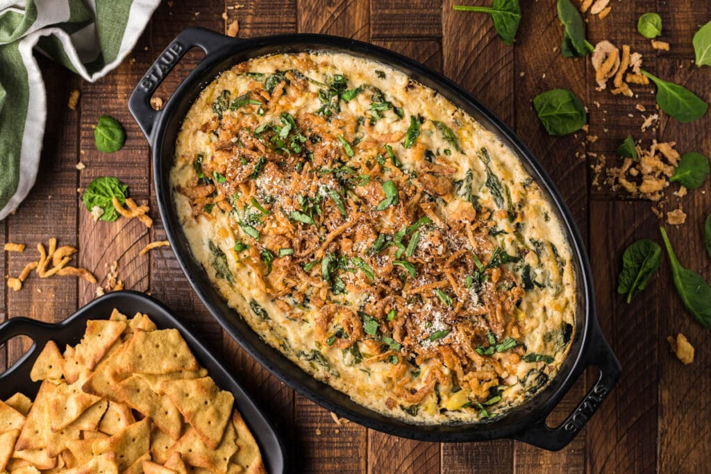 Overhead shot of spinach artichoke dip without mayo in a black oval-shaped baking dish topped with fried onions and cut basil leaves and placed on a wooden table. Black, square-shaped bowl with chips to the left of the baking dish. Spinach leaves and fried onion pieces spread around the table.