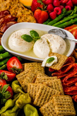 Closeup photo of the Burrata Cheese Board. A white container with Burrata cheeses is surrounded by various ingredients.