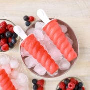 Two red Kool-Aid Popsicles placed on top of a bowl full of ice and surrounded by fresh berries.