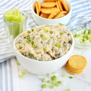 A bowl of buffalo chicken spinach dip on the table with a bowl of crackers and celery sticks.