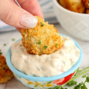 An air fryer catfish nugget held by fingers dipping into a bowl of tartar sauce.
