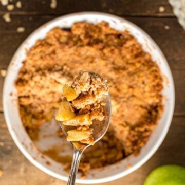 A spoon lifting a serving of apple crumble from a bowl, with whole apples and a rustic wooden table in the background.