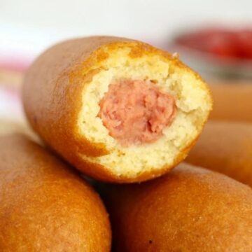 A close-up image of a pile of corn dogs, one with a bite taken out revealing the hot dog inside, with condiments blurred in the background.
