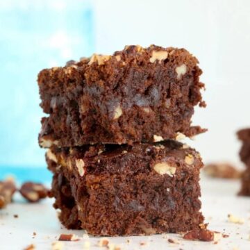 Stack of three fudgy brownies with pecans on a white surface, with more pecans scattered around and a blue glass in the background.