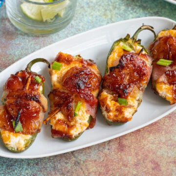 A plate of bacon-wrapped jalapeño peppers garnished with green onions, served on a white oval plate with a blue napkin and a drink in the background.