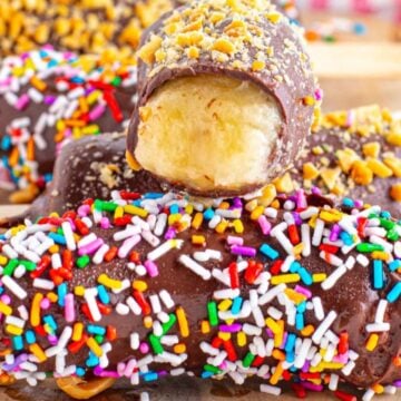 Close-up of chocolate-coated frozen banana on sticks, covered with colorful sprinkles and chopped nuts, displayed on a wooden board.