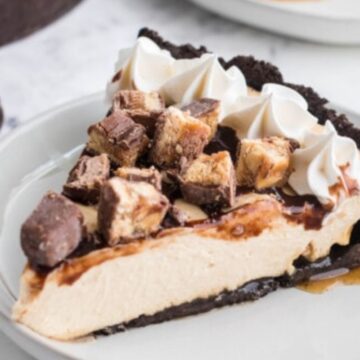 A slice of creamy chocolate cheesecake with cookie crumble topping and whipped cream on a white plate, with whole cookies in the background.