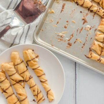 Twisted pastry sticks made from refrigerated dough, drizzled with chocolate on a baking sheet and a white plate, accompanied by a metal spatula and a striped cloth on a light wooden surface.