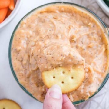 A hand holding a cracker dipped in creamy dip, with bowls of carrots and cucumber slices along with other party dips in the background.