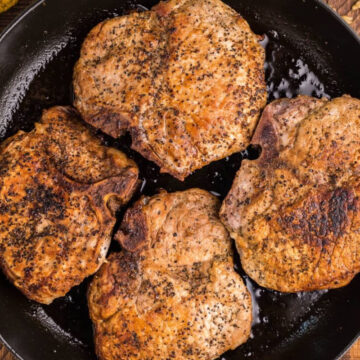 Four seasoned and cooked pork chops in a black skillet with a green handle, on a wooden surface. This easy pork chop recipe is perfect for family meals.