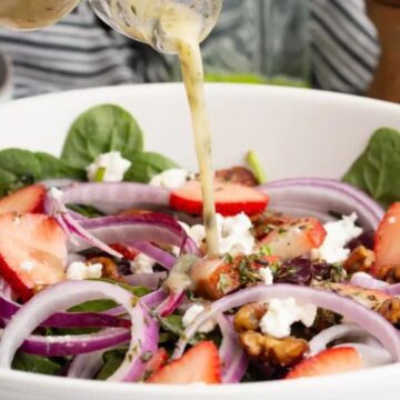 A fresh spinach salad with strawberry slices, red onions, feta cheese, walnuts, and a creamy dressing being poured over.