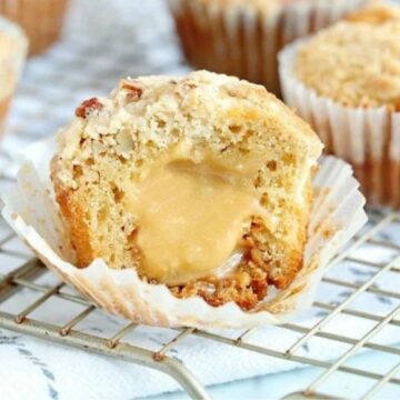 A muffin with a gooey caramel center on a cooling rack, surrounded by coffee beans, with other whole muffins blurred in the background.