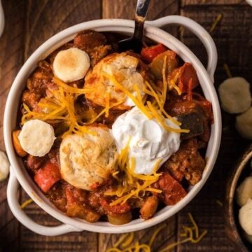 A bowl of chili topped with shredded cheese, sour cream, and oyster crackers, surrounded by ingredients including Pillsbury biscuits on a wooden table.