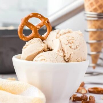 A bowl of homemade peanut butter ice cream garnished with a pretzel, with scattered peanuts and a waffle cone in the background.