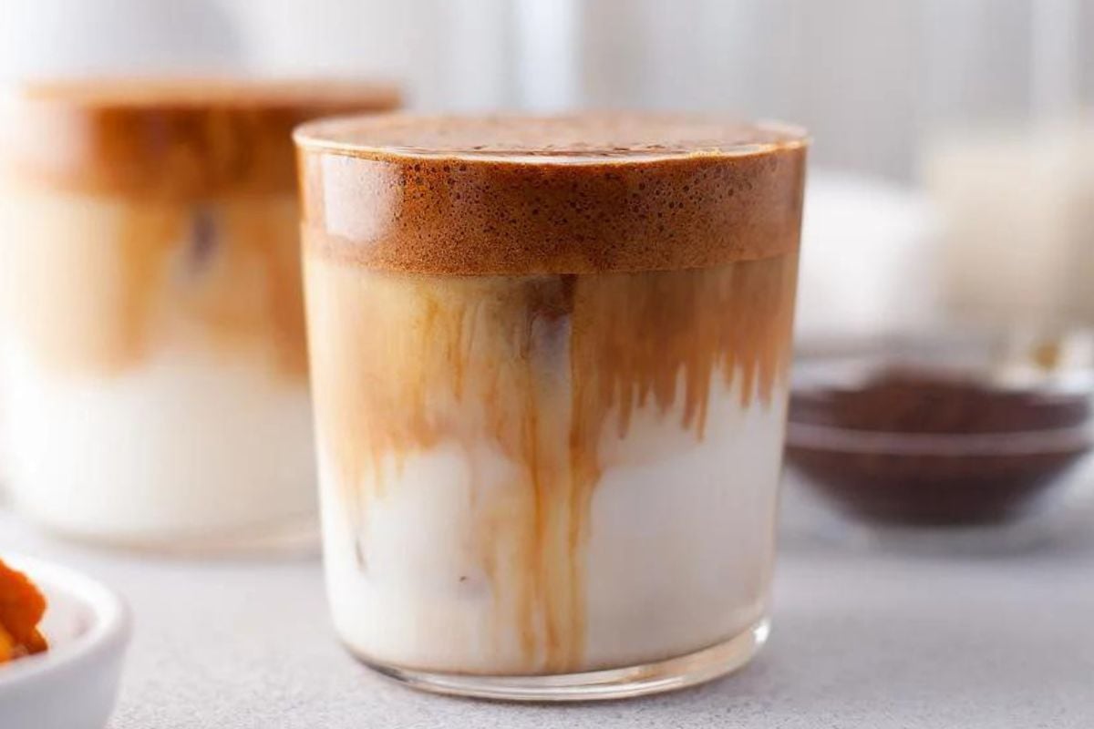 A glass of iced dalgona coffee with a frothy whipped coffee top layer above milk, showing swirls where the coffee mixes with the milk in a cozy coffee shop.