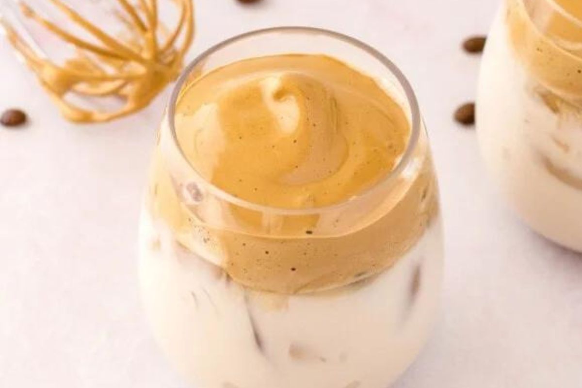 A glass of dalgona coffee, featuring a creamy whipped coffee topping over milk, garnished with a few coffee beans on the side.