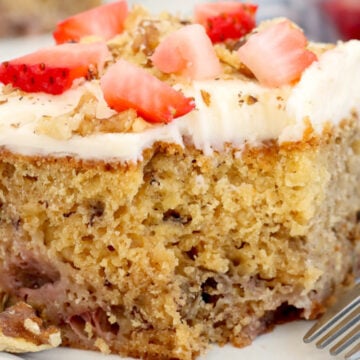 A slice of banana cake topped with fresh strawberries and frosting, garnished with nuts on a white plate, with a fork on the side.