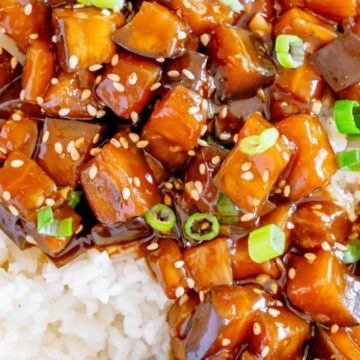 A close-up of sweet and sour tofu served over white rice, garnished with sesame seeds and chopped green onions alongside vegetable sides.