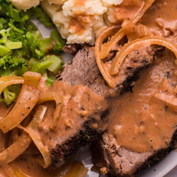 A plate of slow cooker roast beef with gravy and onions, served with mashed potatoes and steamed broccoli.