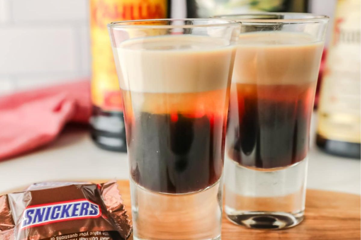 Two layered shots of B-52 cocktails in clear glasses on a wooden surface, with a Snickers bar and liquor bottles in the background&mdash;perfect for a late-night snack.