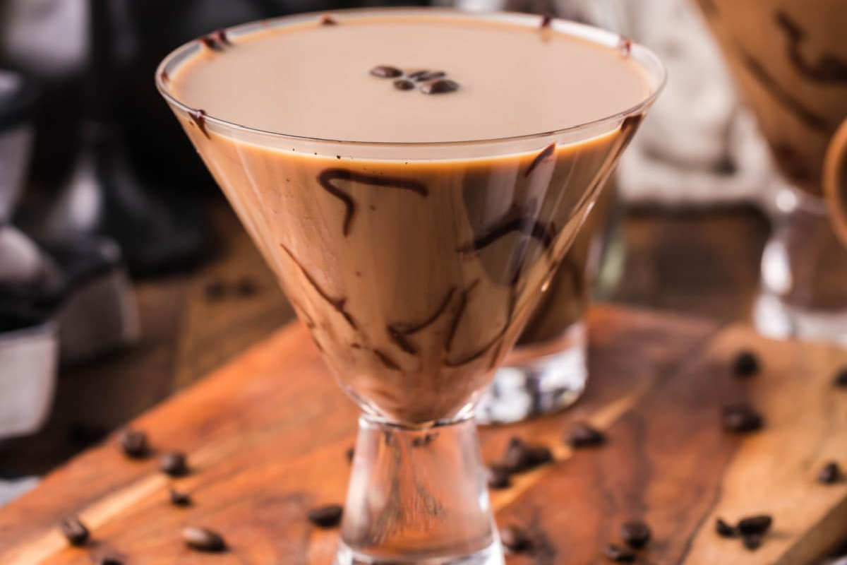 A martini glass filled with a creamy, coffee-colored alcoholic drink, garnished with coffee beans on top and chocolate drizzles on the glass, placed on a wooden surface with coffee beans scattered around.