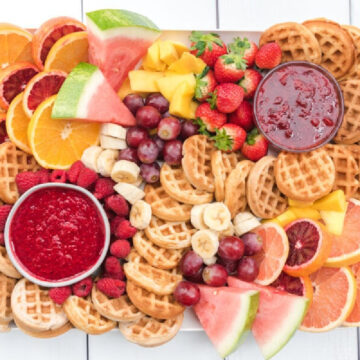 A large platter featuring mini waffles, fresh fruits (orange slices, watermelon, mango, strawberries, grapes, raspberries, and bananas), and two bowls of red fruit spreads—perfect for a delightful brunch or breakfast.