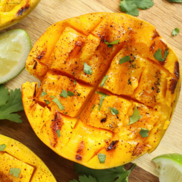 Grilled mango halves garnished with lime wedges and cilantro leaves on a wooden surface, perfect as refreshing side dishes.