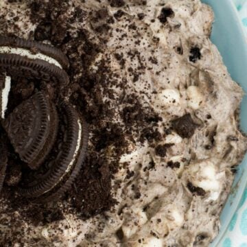 Close-up of a cookies and cream dessert made with Oreo cookies, featuring crushed and whole chocolate sandwich cookies, sitting on a light blue surface with blue and white patterned fabric beside it. This is one of those easy treats perfect for any occasion.