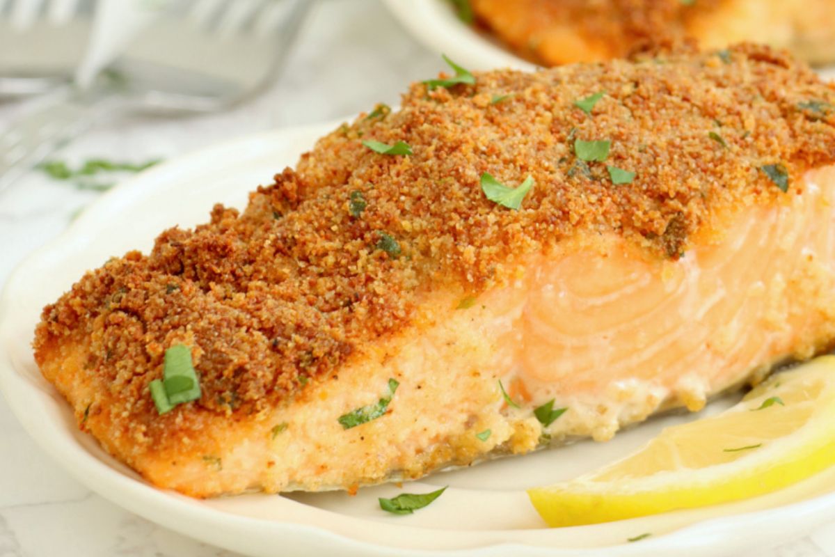 A close-up of a baked salmon fillet topped with a crispy breadcrumb crust, garnished with chopped herbs, and served with a lemon wedge on a white plate.
