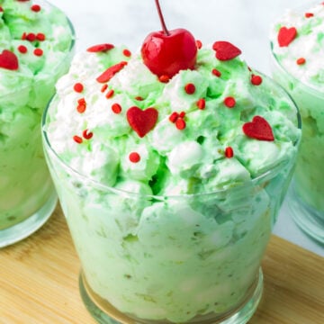 Three glass bowls filled with Fluff salad, topped with red heart-shaped sprinkles, red round sprinkles, and a cherry, are placed on a wooden surface.