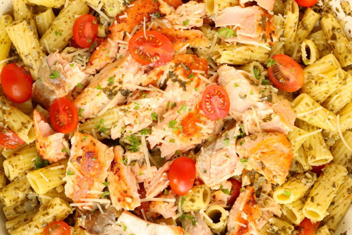 A close-up of a pasta dish featuring rigatoni with pesto, topped with cooked salmon pieces, cherry tomatoes, and grated cheese.