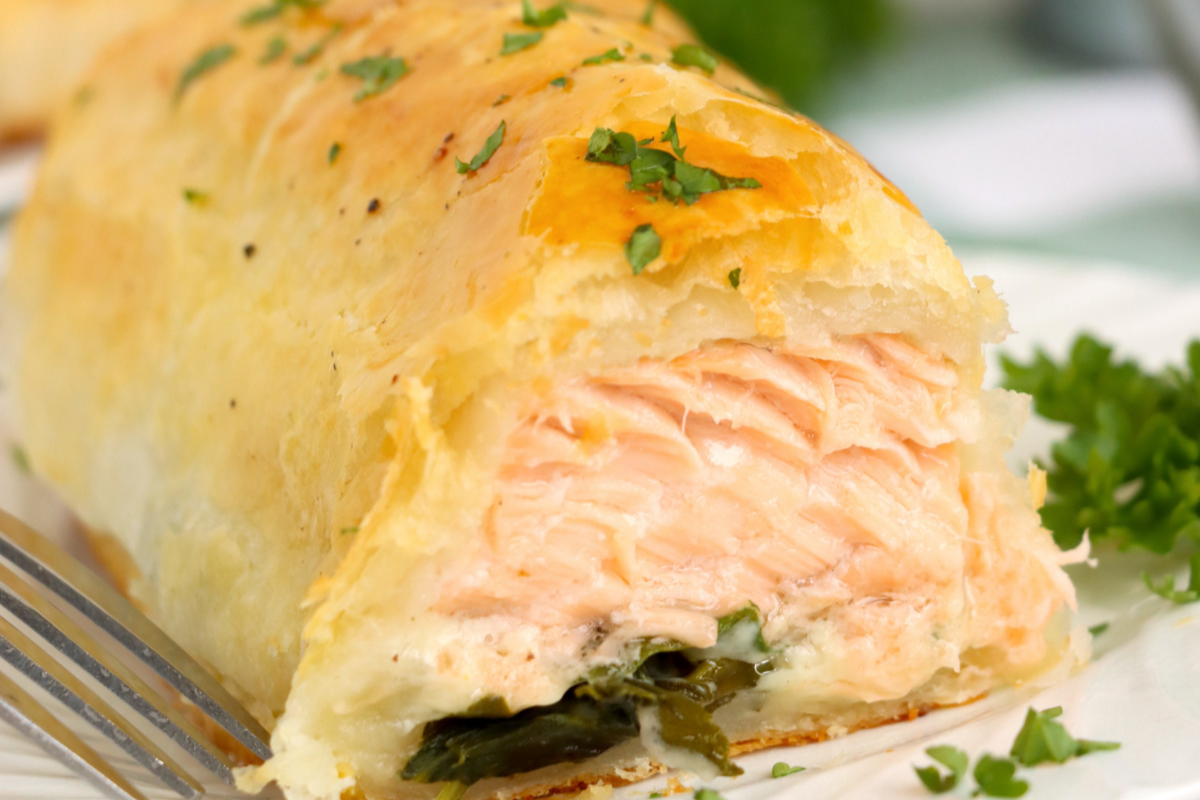 Close-up of a partially sliced salmon Wellington with golden puff pastry, filled with spinach and a creamy mixture. The dish is garnished with chopped parsley. A fork is placed beside it.