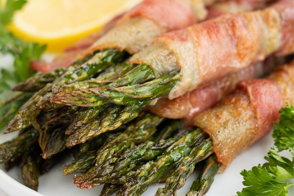 Close-up of bacon-wrapped asparagus spears on a white plate, spotlighting the vibrant green and crispy bacon, garnished with fresh parsley and a lemon slice in the background. Ideal as one of your standout side dishes.